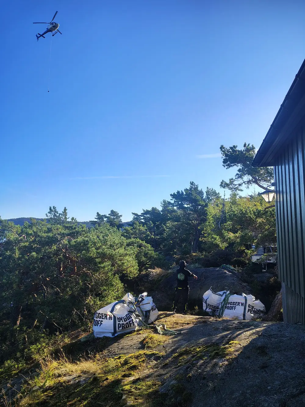 Sekker med materialer på svaberget mellom furutrær, med hytta i bakgrunnen