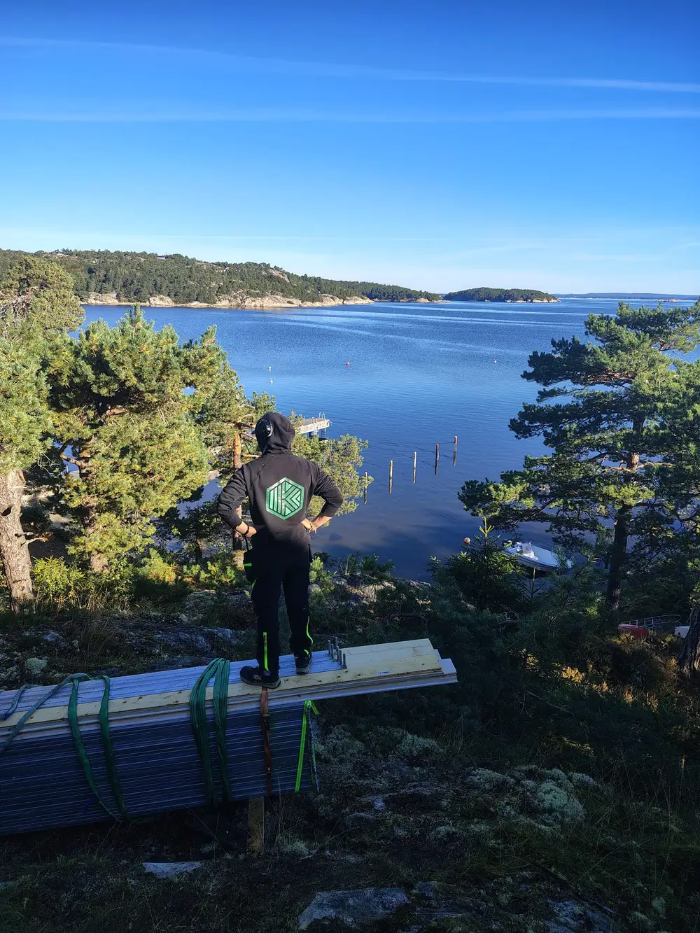 Tømrer med ryggen til ser ut over sjøen fra svaberget ved hytta, med materialer ved føttene