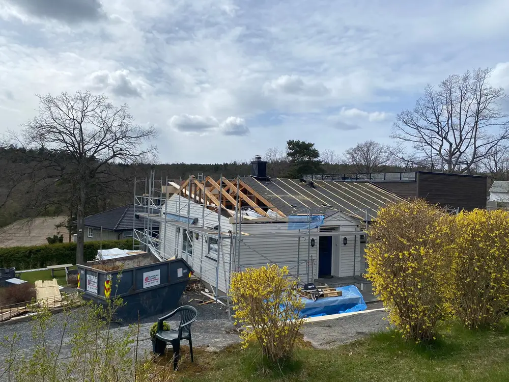 Hus med nye takstoler i lyst treverk, stillas langs veggen og en container foran